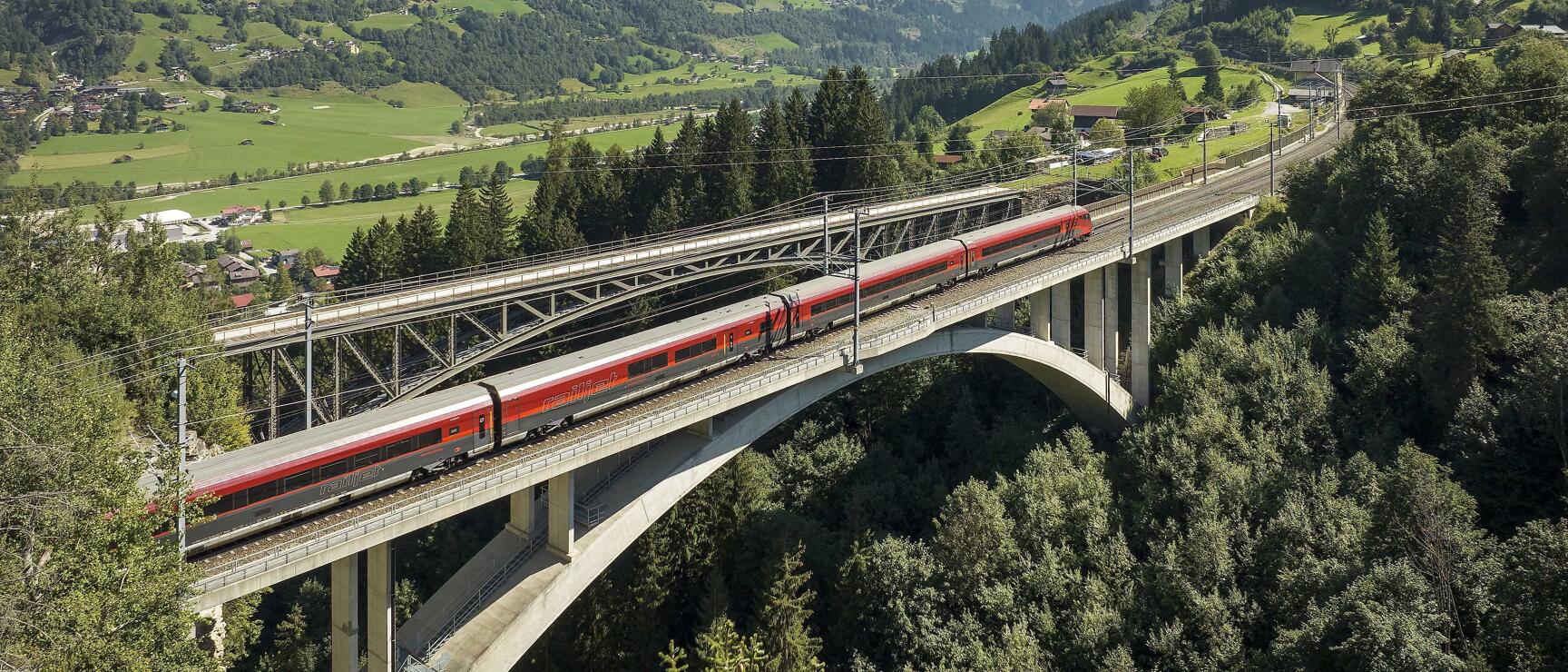 Roter ÖBB-Zug auf Betonbrücke über bewaldeter Schlucht, grünes Alpental mit Bergen im Hintergrund.