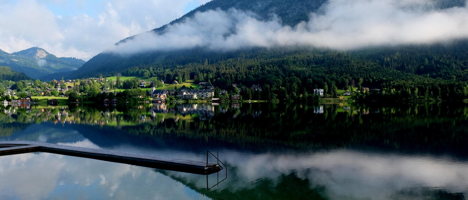 Wooden dock on a calm mountain lake, village and forested mountain with mist reflected in water