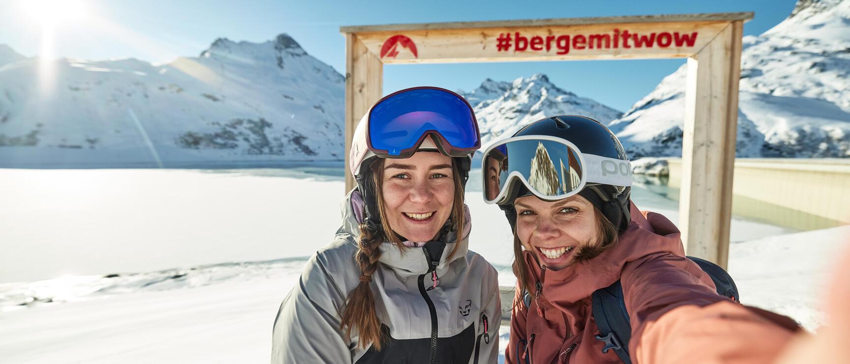 Two skiers wearing helmets and goggles in front of a wooden frame with '#bergemitwow', snow-covered mountains in the background.