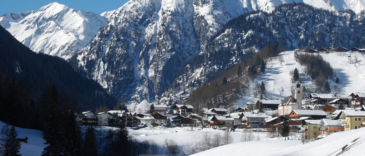 Winter alpine village with church, snow-covered houses and steep mountain peaks in the background.
