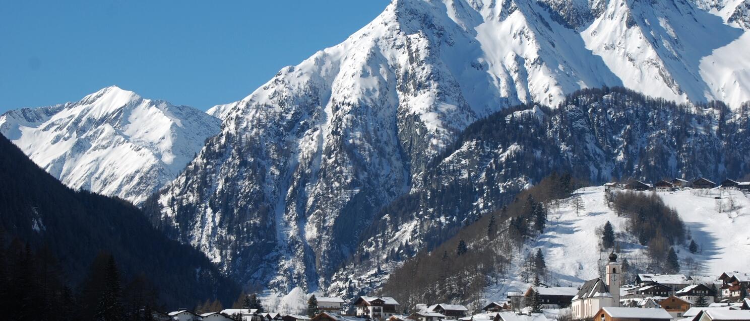 Winter alpine village with church, snow-covered houses and steep mountain peaks in the background.