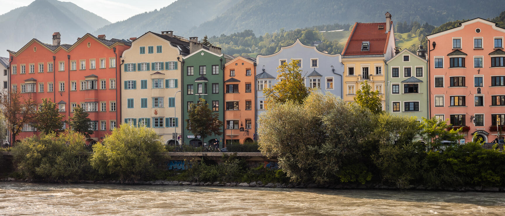 Colorful row of houses on the Inn riverbank in Innsbruck, forested mountains in the background under cloudy sky.