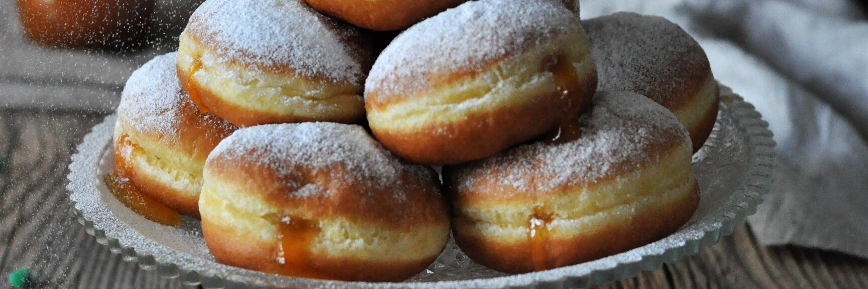 Multiple donuts with powdered sugar on a glass stand, bitten donut with jam in front, colorful confetti.