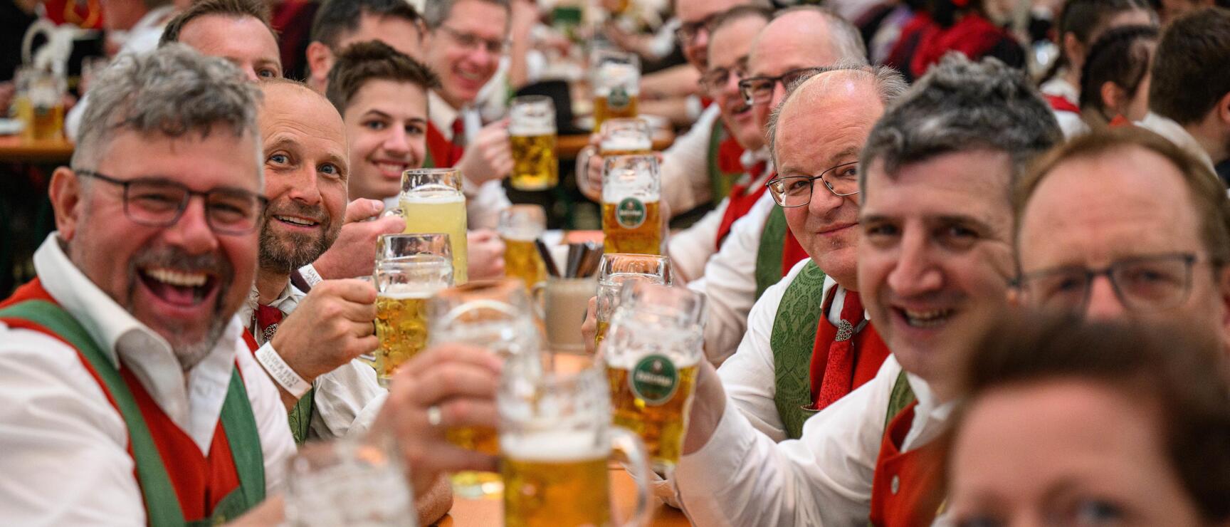 Men in traditional clothing raising beer mugs at long table in crowded beer tent.