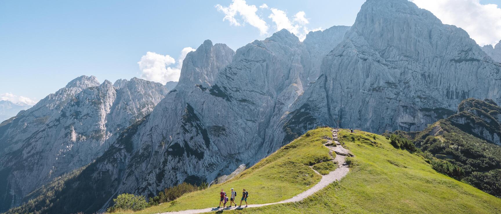 Drei Wanderer auf Gratweg mit grüner Wiese, dahinter markante Felswände unter blauem Himmel.