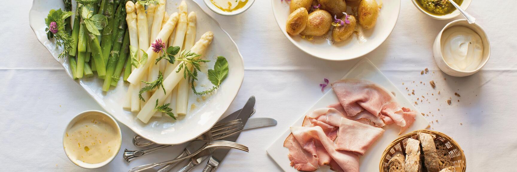 Plate with green and white asparagus, potatoes, ham, bread and sauces on white tablecloth.