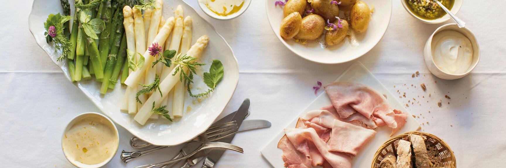 Teller mit grünem und weißem Spargel, Kartoffeln, Schinken, Brot und Saucen auf weißem Tischtuch.