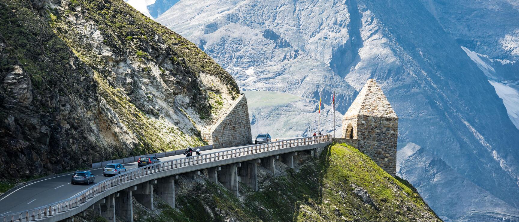 Cars on Großglockner High Alpine Road with stone tower and snow-capped peaks in the background.