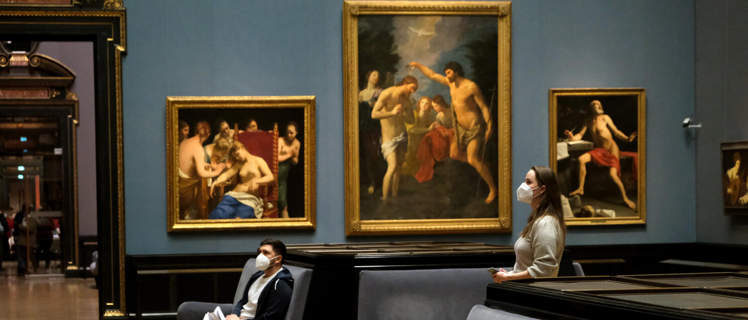 Two people wearing masks in a museum hall with large paintings in gold frames, Vienna.
