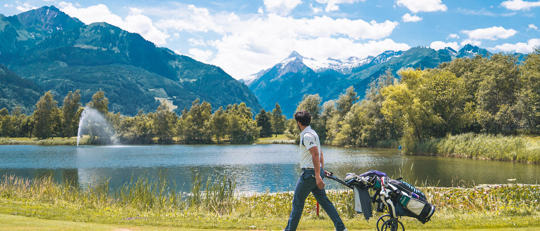 Golfer with trolley on a golf course, lake with fountain and snow-capped mountain peaks in the background.