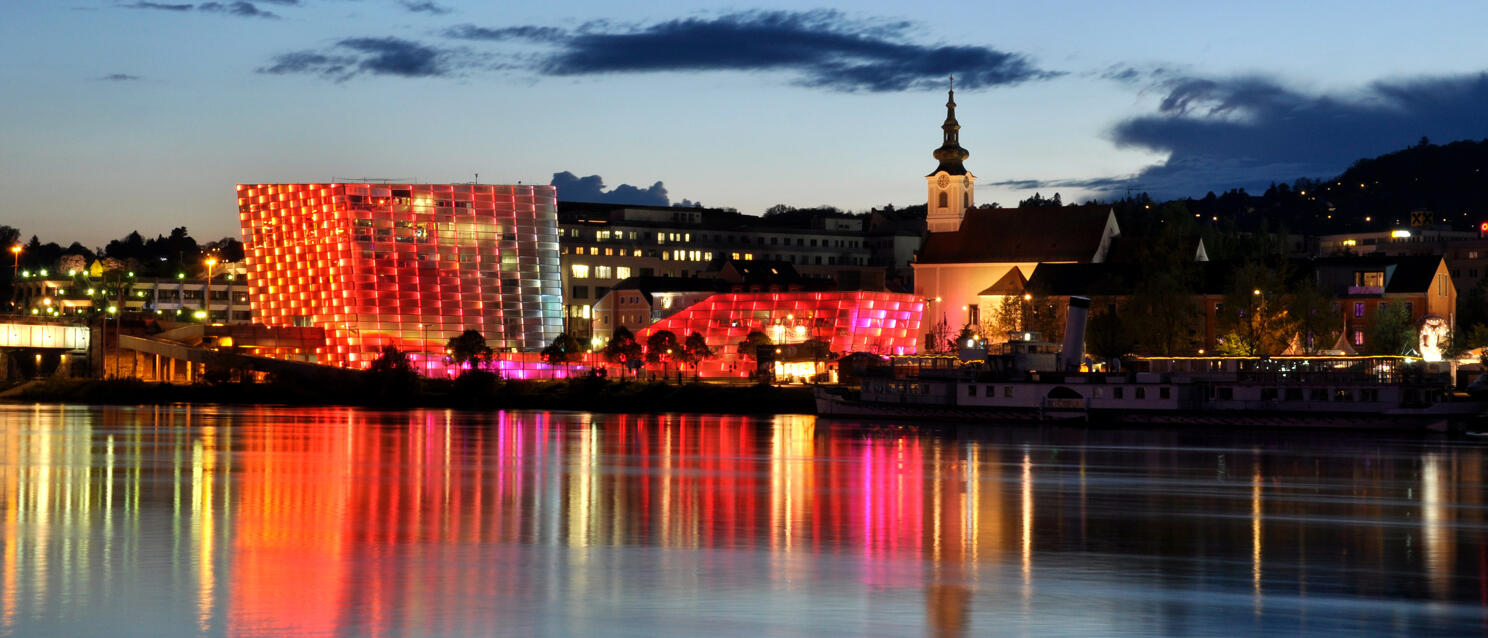 Red-lit Ars Electronica Center reflected in the Danube, Linz church tower and city skyline in background.