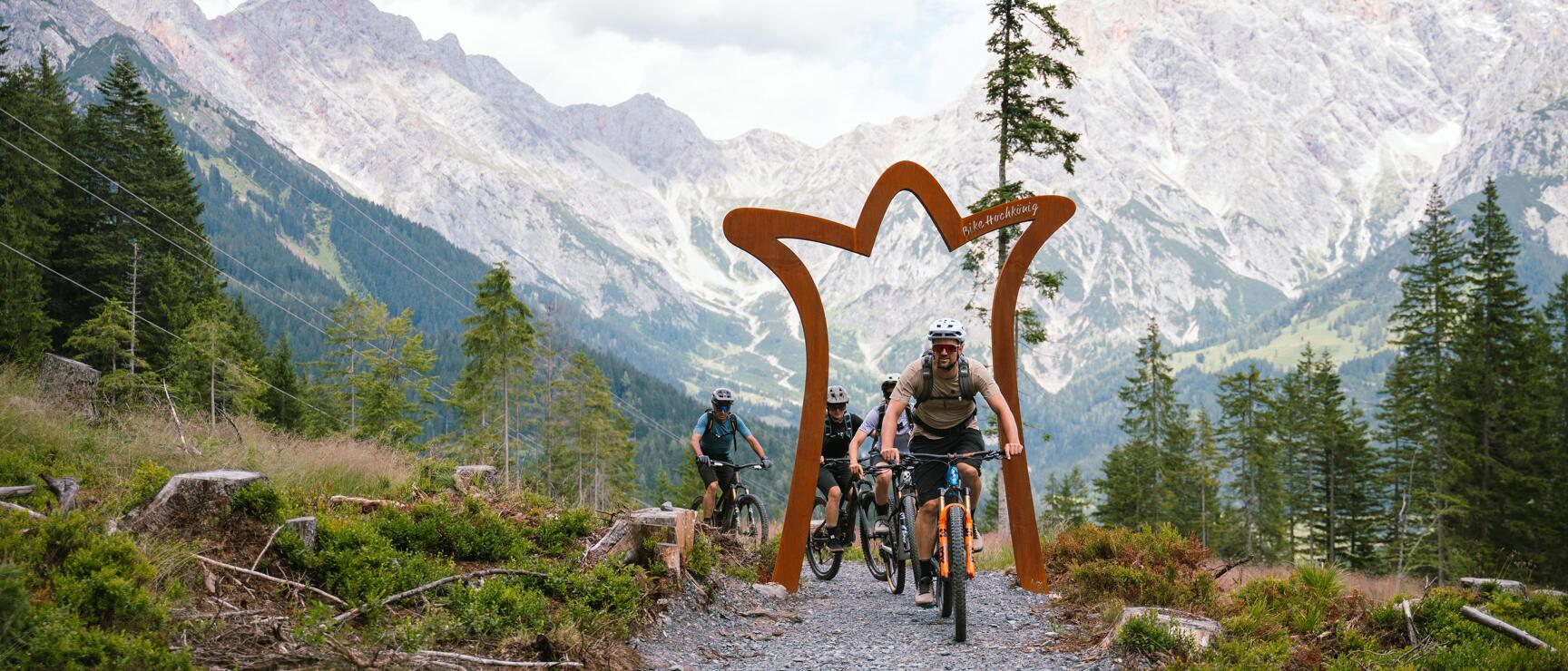 Drei Mountainbiker fahren unter einem Bike-Hochkönig-Stahltor auf einem Schotterweg, Bergpanorama im Hintergrund.
