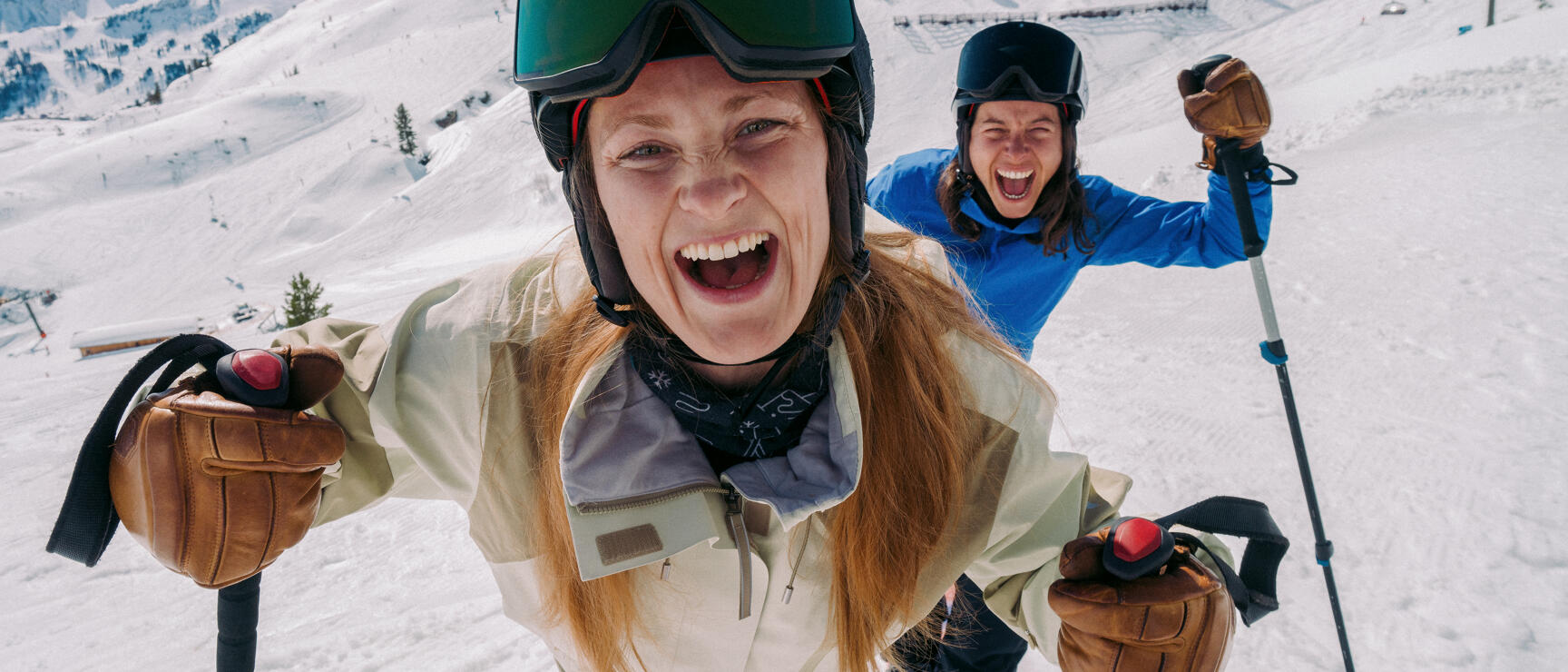 Two laughing female skiers with helmets and ski goggles on a snowy slope, mountain panorama in the background.