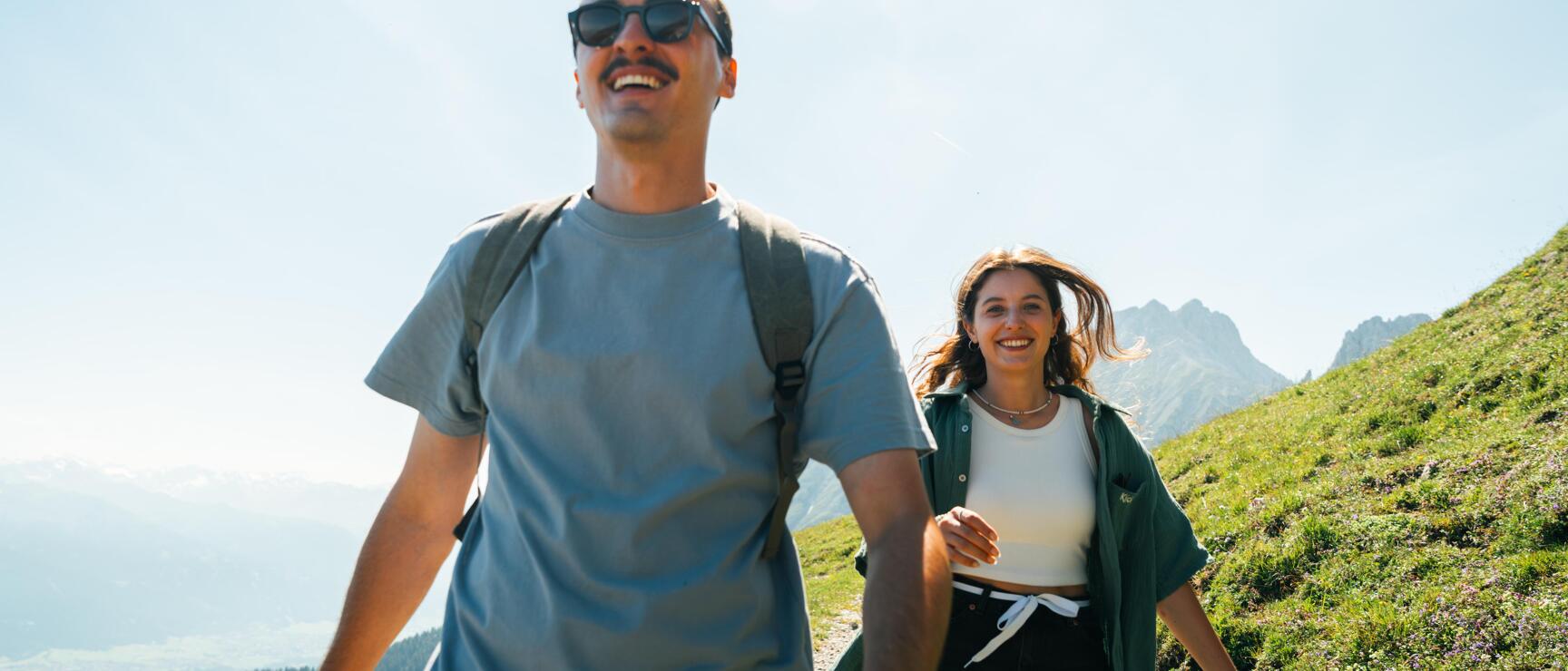 Two people are hiking on the Nordkette in Innsbruck in sunny weather. In the foreground, a person wearing sunglasses and carrying a rucksack is walking, followed by another person smiling. In the background, green slopes and a mountainous landscape can be seen under a clear sky.