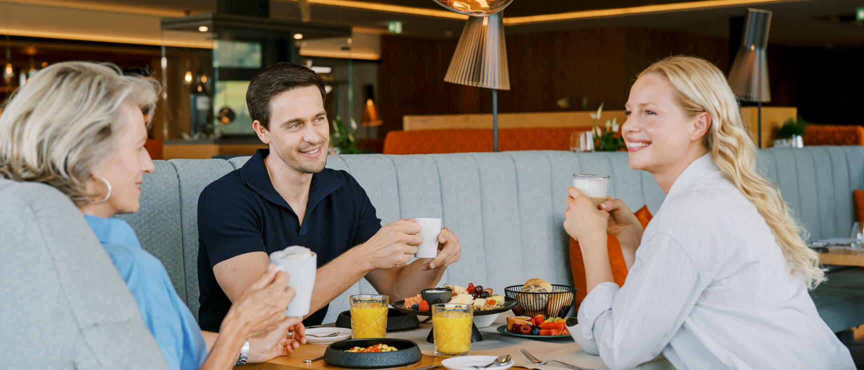 Three people having breakfast in a modern hotel restaurant with coffee cups, orange juice and fruit on the table.