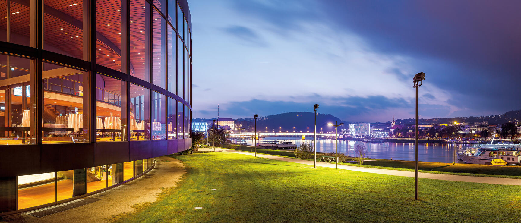 Glass facade of modern building at night, Danube riverside with illuminated bridge and city lights, Linz.