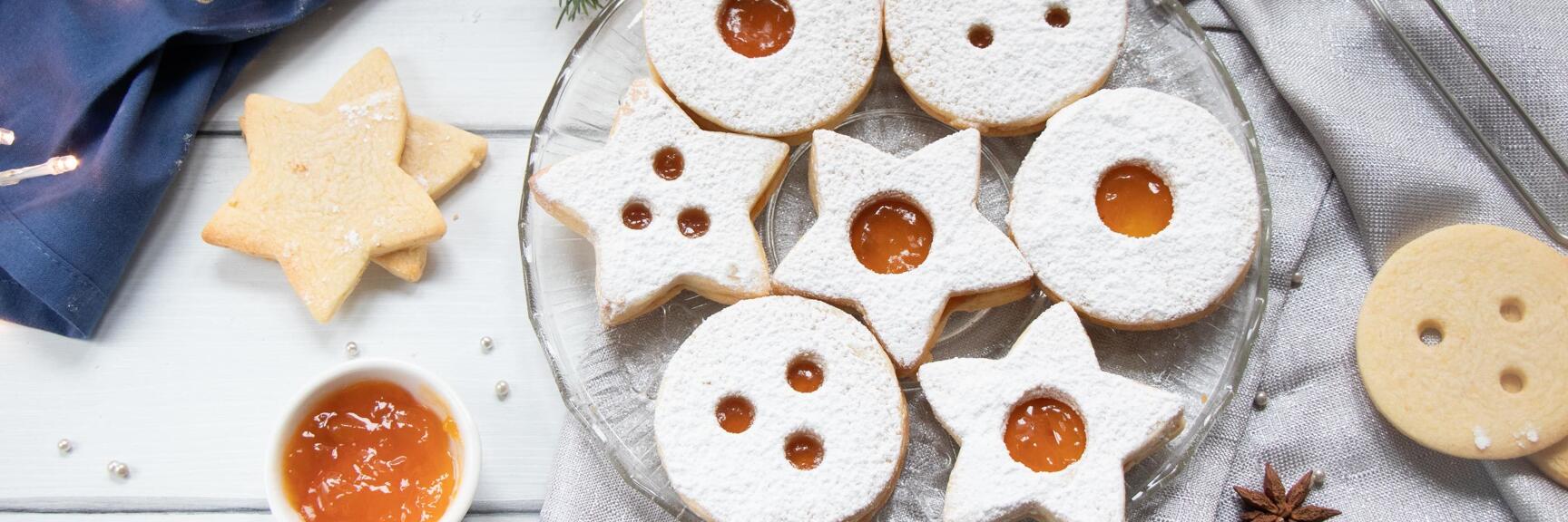 Linzer Augen mit Marillenmarmelade und Puderzucker auf Glasteller, daneben Sieb, Kerze und Tannenzweig.