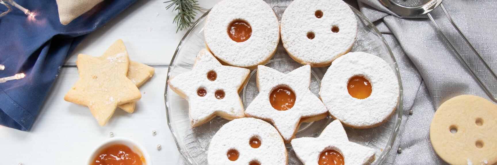 Linzer eyes with apricot jam and powdered sugar on glass plate, beside sieve, candle and fir branch.