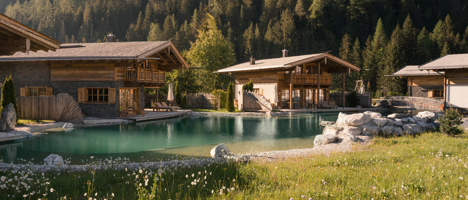 Wooden chalets with natural pool, coniferous forest and rocky peak in background, flower meadow in foreground.