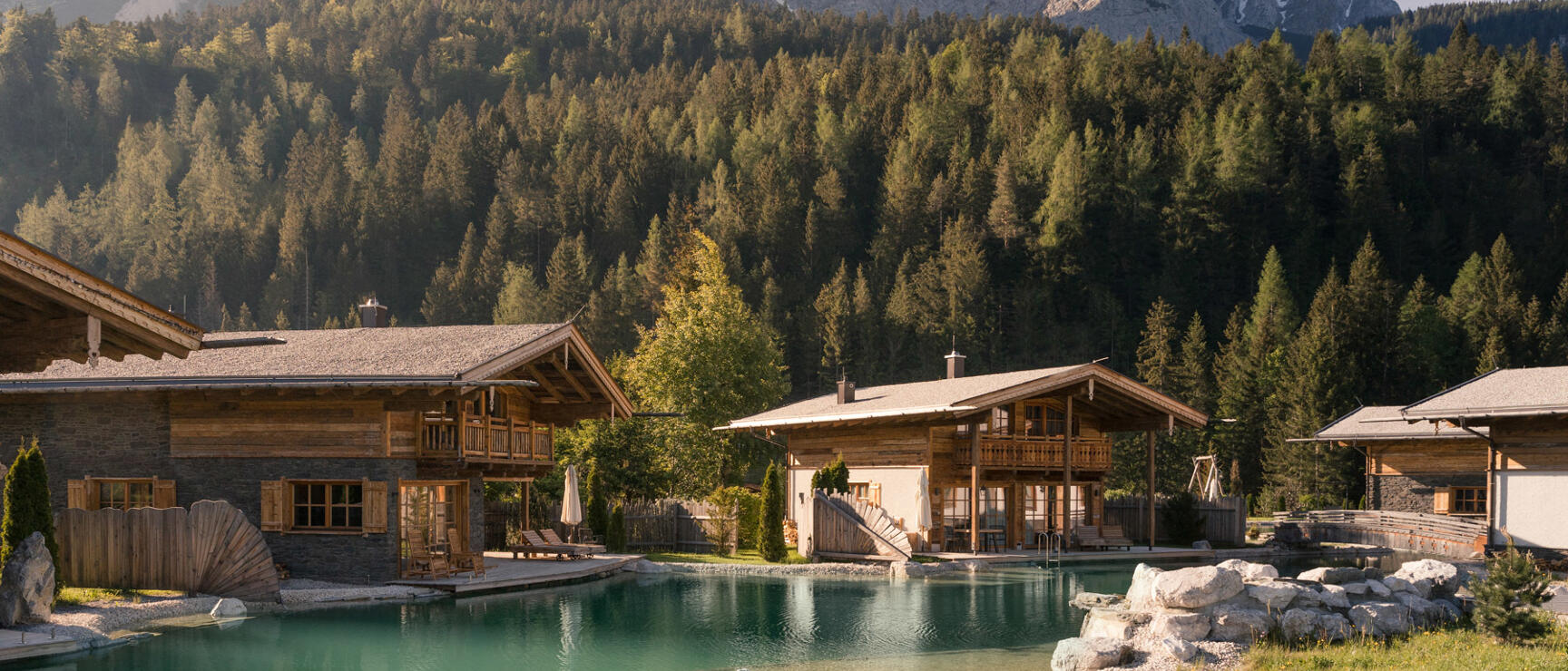 Wooden chalets with natural pool, coniferous forest and rocky peak in background, flower meadow in foreground.