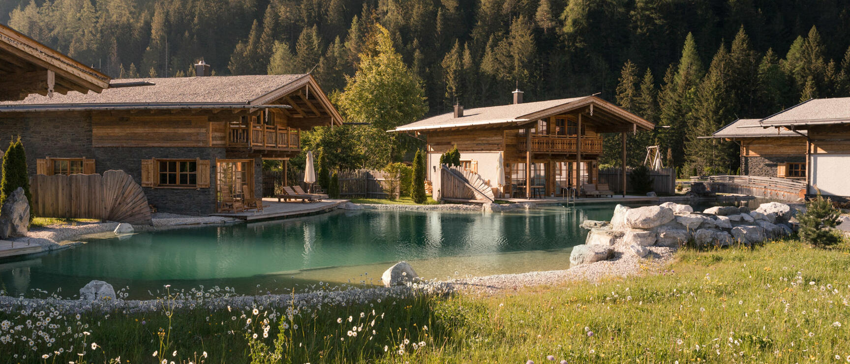 Wooden chalets with natural pool, coniferous forest and rocky peak in background, flower meadow in foreground.