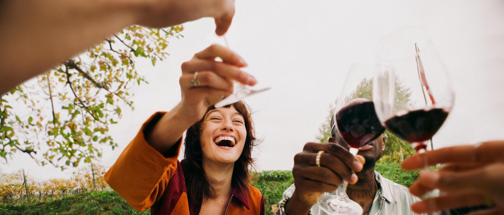 Two people toasting with red wine at a set table outdoors, vineyard in the background.