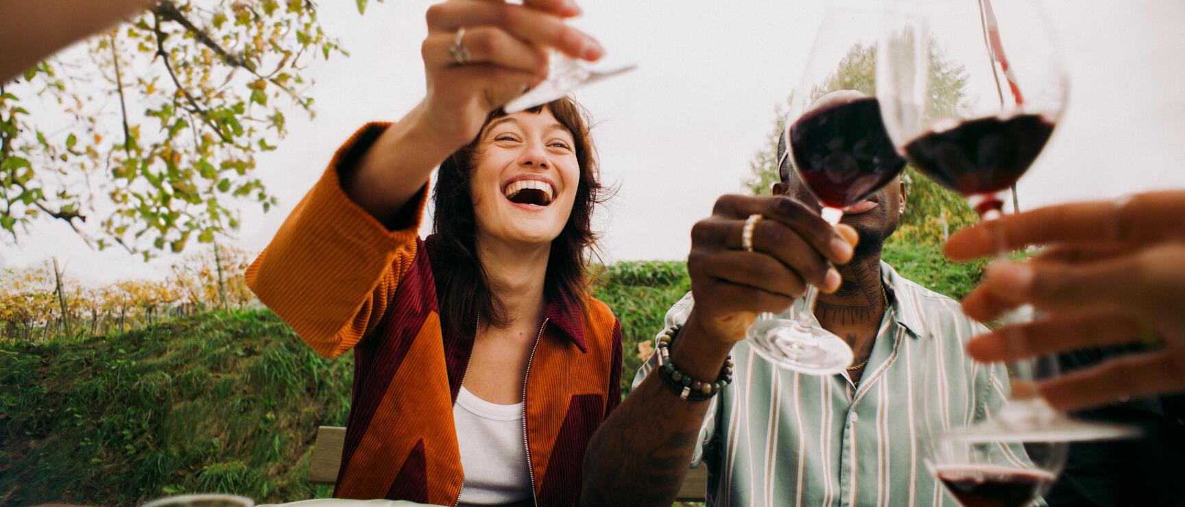 Zwei Personen stoßen mit Rotwein an einem gedeckten Tisch im Freien an, Weinberg im Hintergrund.
