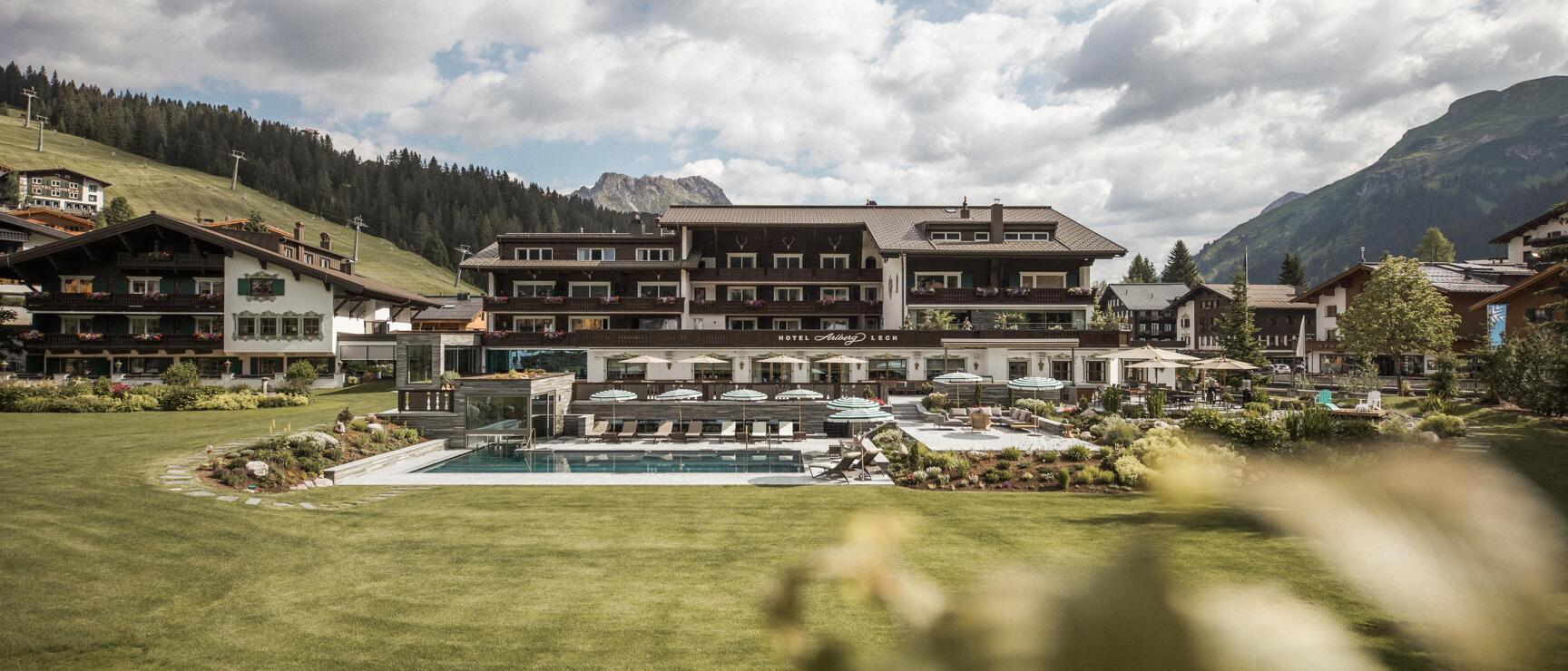Hotel Arlberg Lech with outdoor pool, green meadow and mountain panorama in summer.