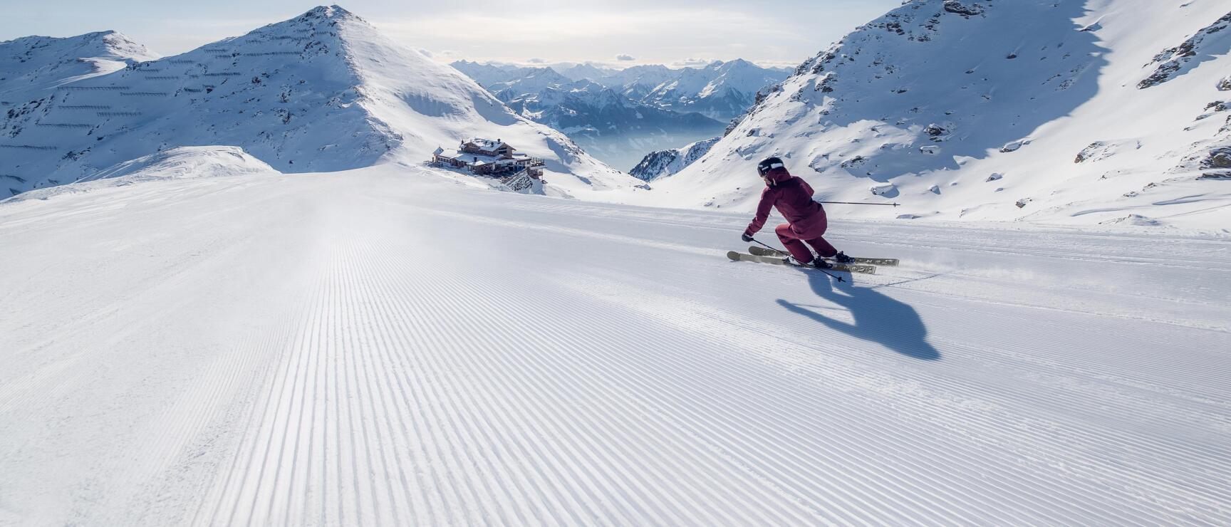 Skier in red suit on prepared slope with snow-covered mountain peaks and sun in background.