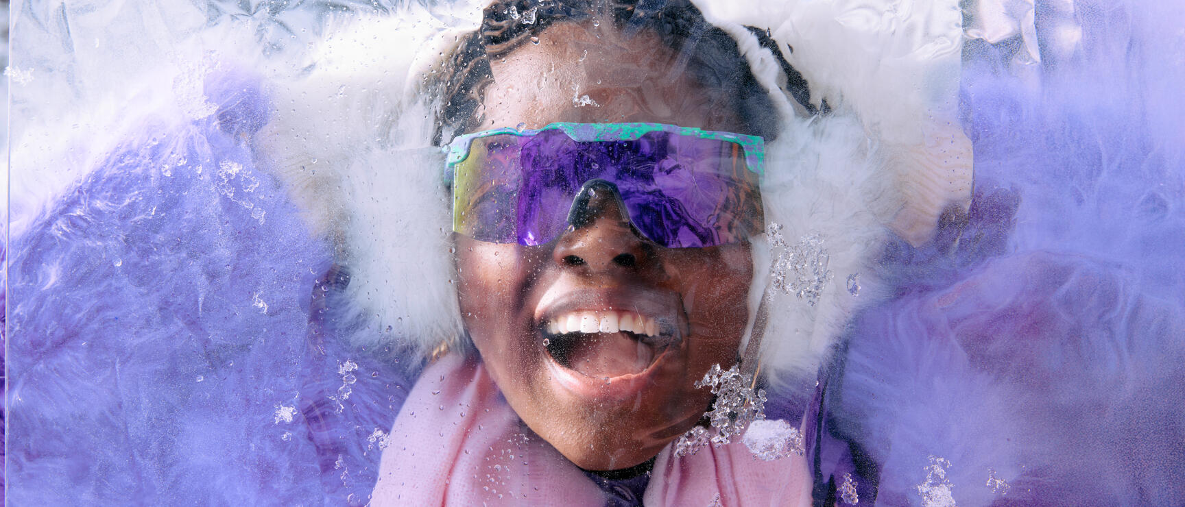 Laughing person with mirrored ski goggles and white ear warmers behind a wet ice block.