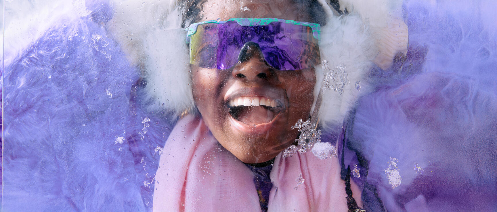 Laughing person with mirrored ski goggles and white ear warmers behind a wet ice block.