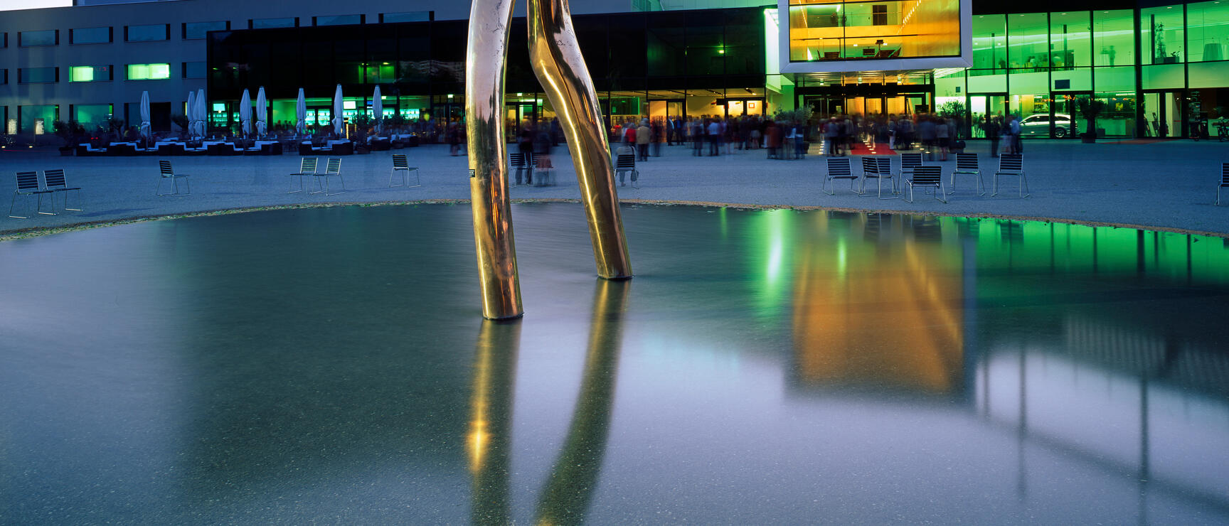 Golden metal sculpture in front of illuminated congress building at dusk, reflected in water basin.