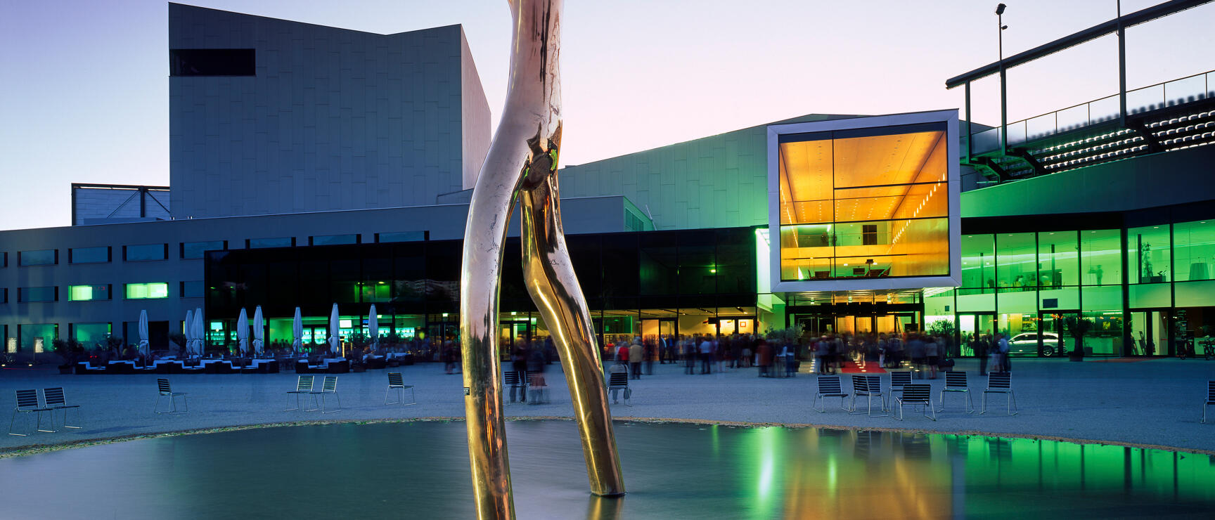 Golden metal sculpture in front of illuminated congress building at dusk, reflected in water basin.