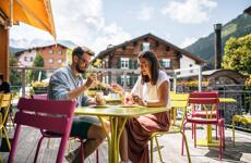 Couple with drinks on colourful café terrace, wooden house and mountain panorama in background.
