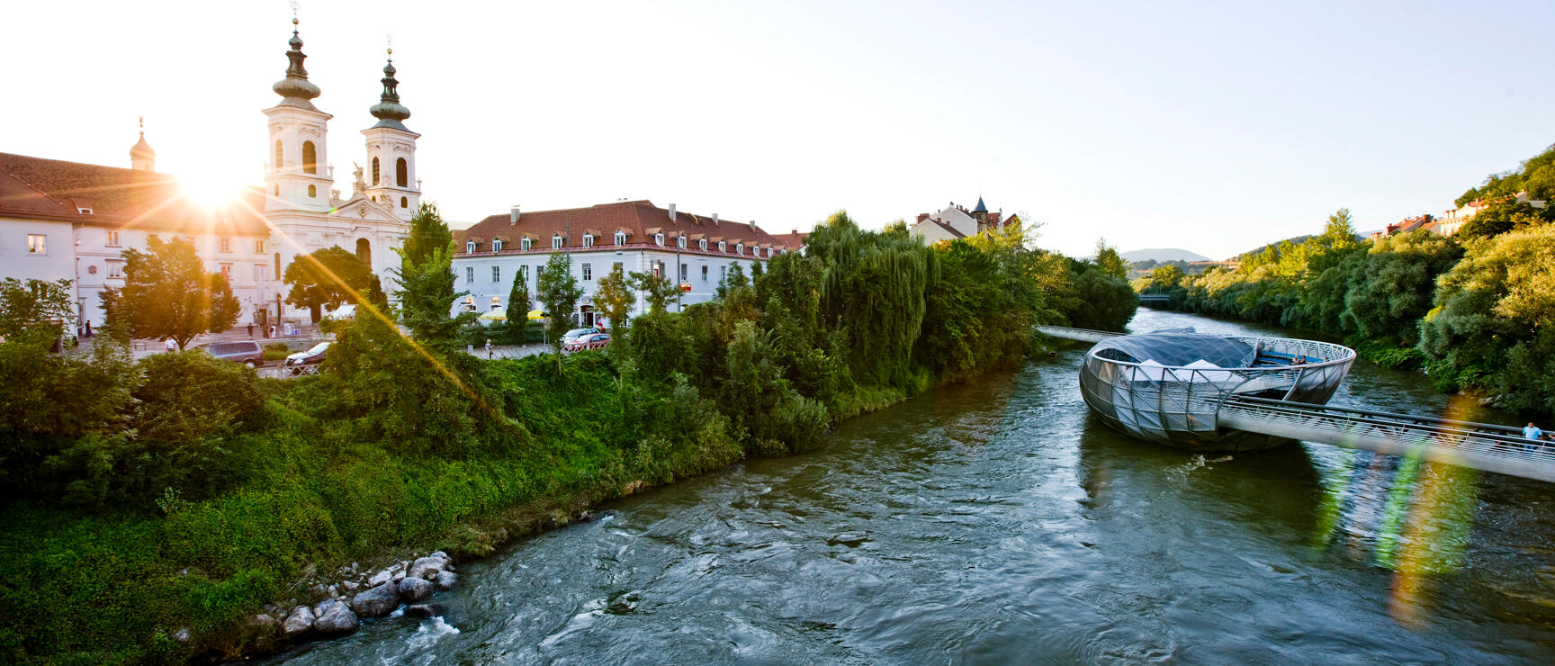 Mur River with Murinsel and Mariahilferkirche church in Graz at sunset, green banks.