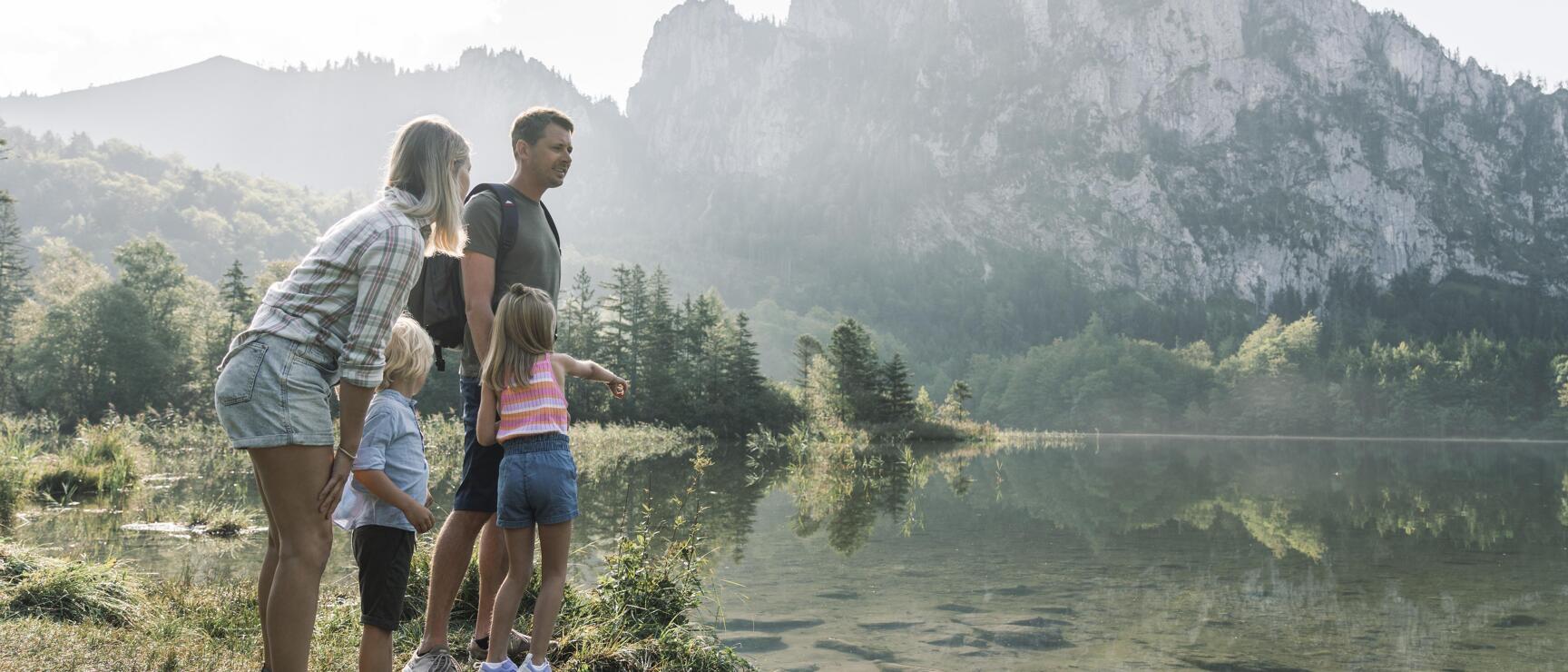 Two adults and two children standing on the shore of a mountain lake with rocky peaks and forest in the background.