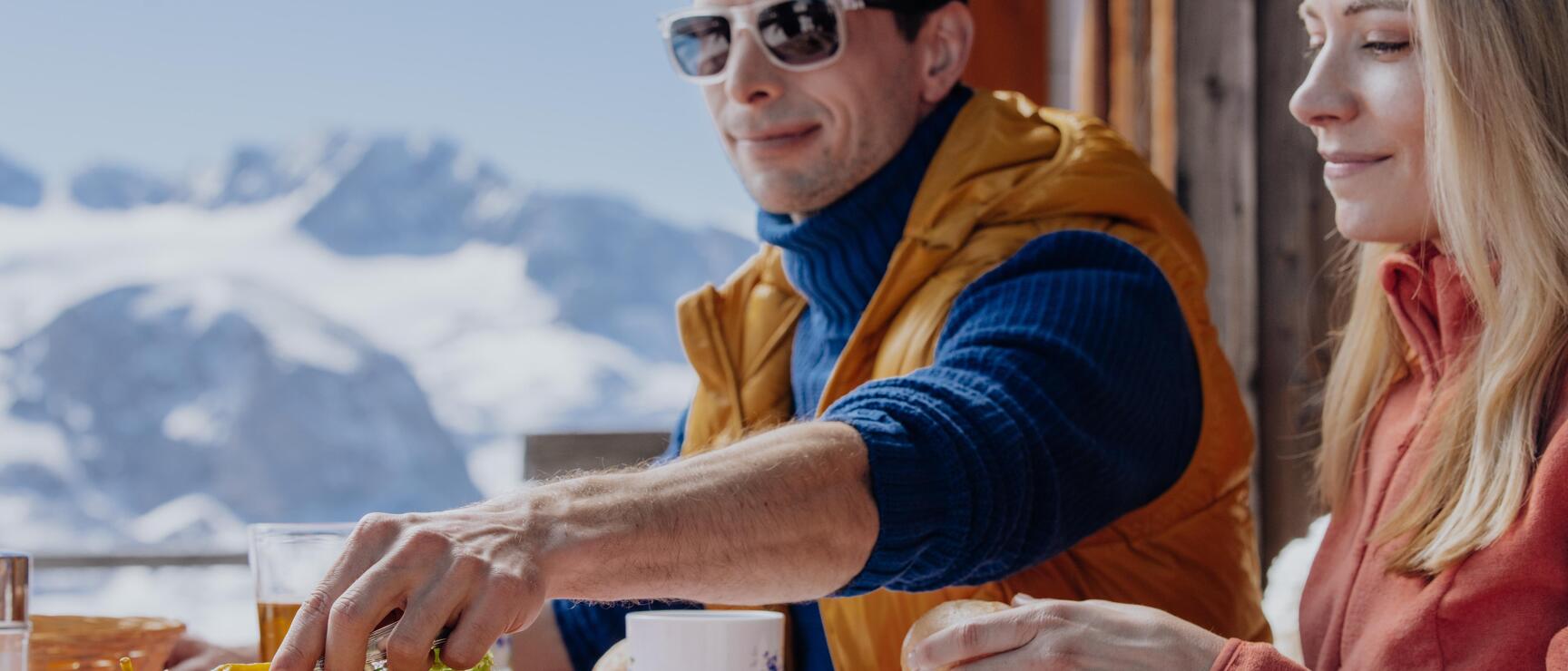 Zwei Personen beim Essen auf einer Almhütten-Terrasse, Brettljause, schneebedeckte Berge im Hintergrund.