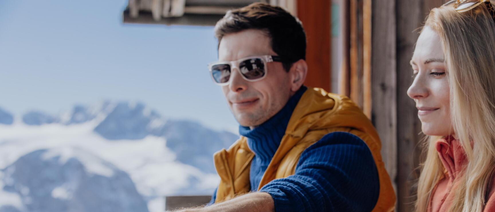 Two people eating on an alpine hut terrace, Brettljause, snow-capped mountains in background.