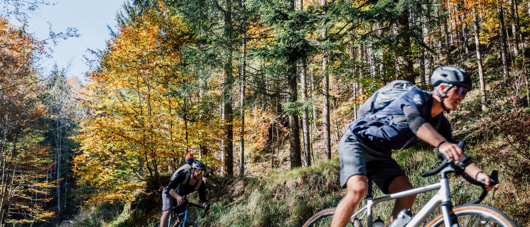 Two cyclists wearing helmets on a gravel path through autumn forest with fallen leaves on the ground.