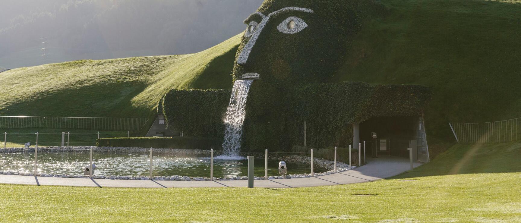 Giant head sculpture with waterfall at Swarovski Crystal Worlds in Wattens, pond and mountain backdrop.
