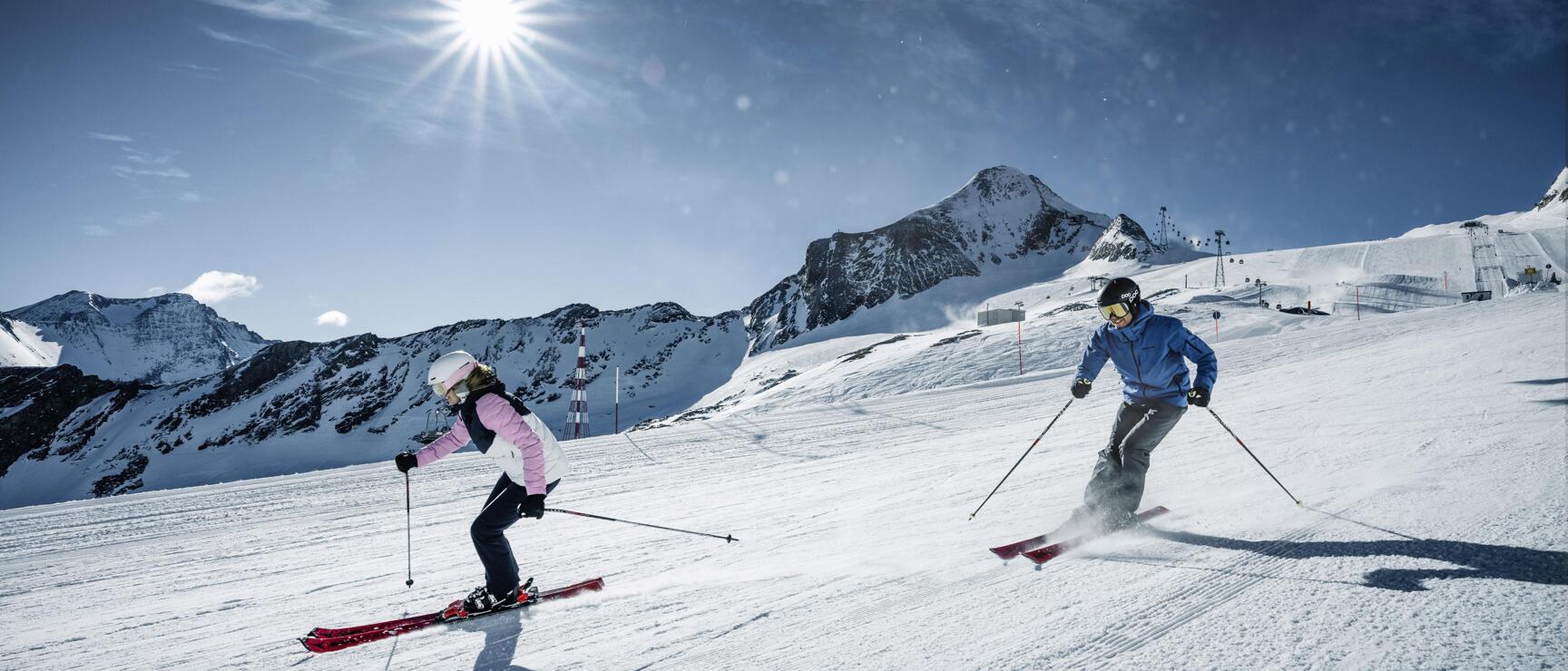 Two skiers on a groomed slope with snow-covered mountain peaks and bright sunshine in the background.