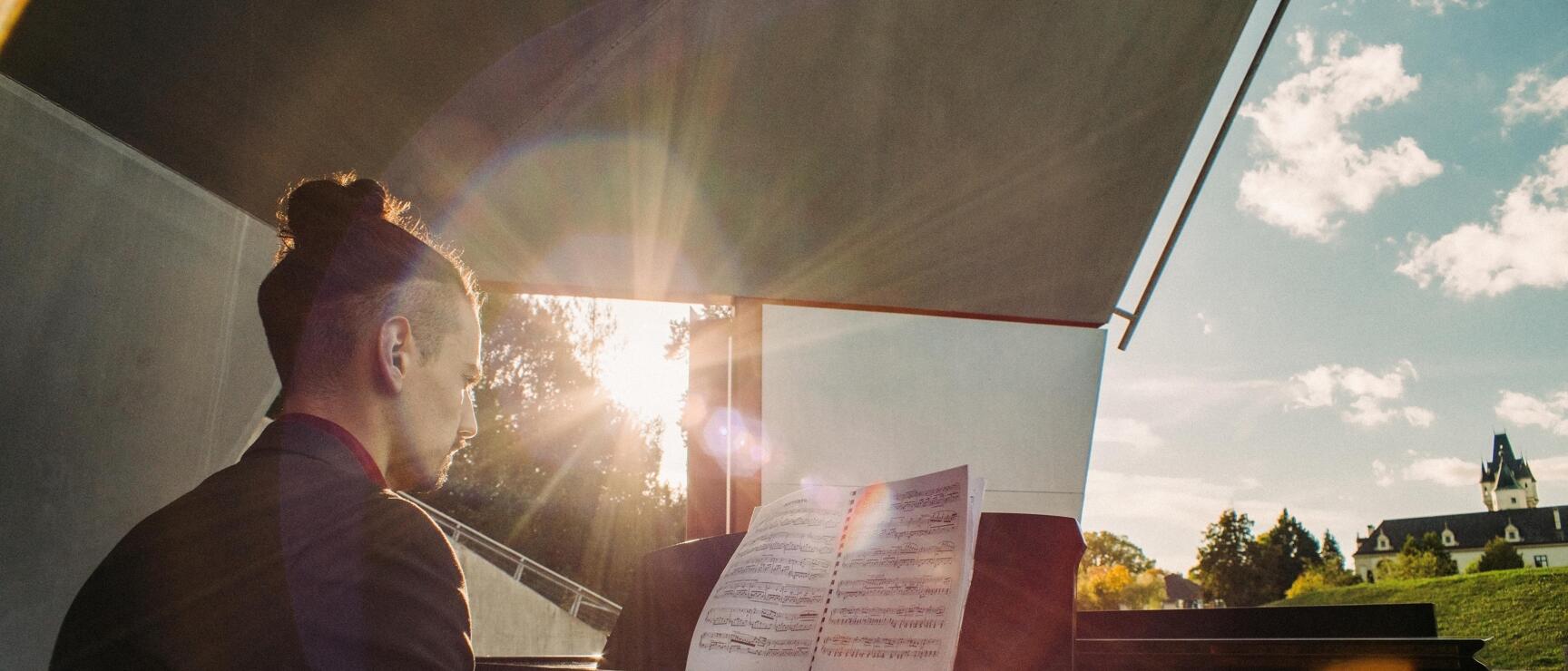 Man playing grand piano outdoors with sheet music, castle and blue sky in background, sunrays