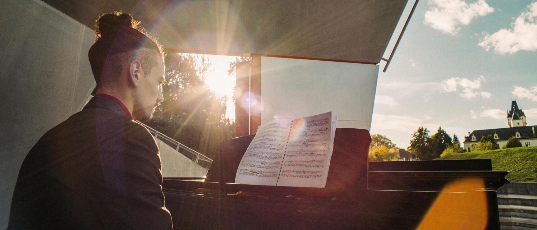 Man playing grand piano outdoors with sheet music, castle and blue sky in background, sunrays