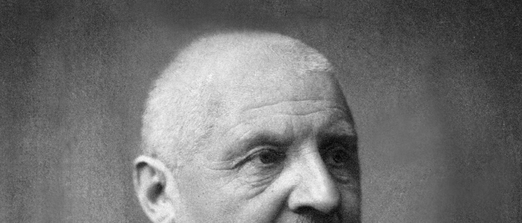 Black and white portrait of an elderly man with a bald head, small moustache and dark jacket.