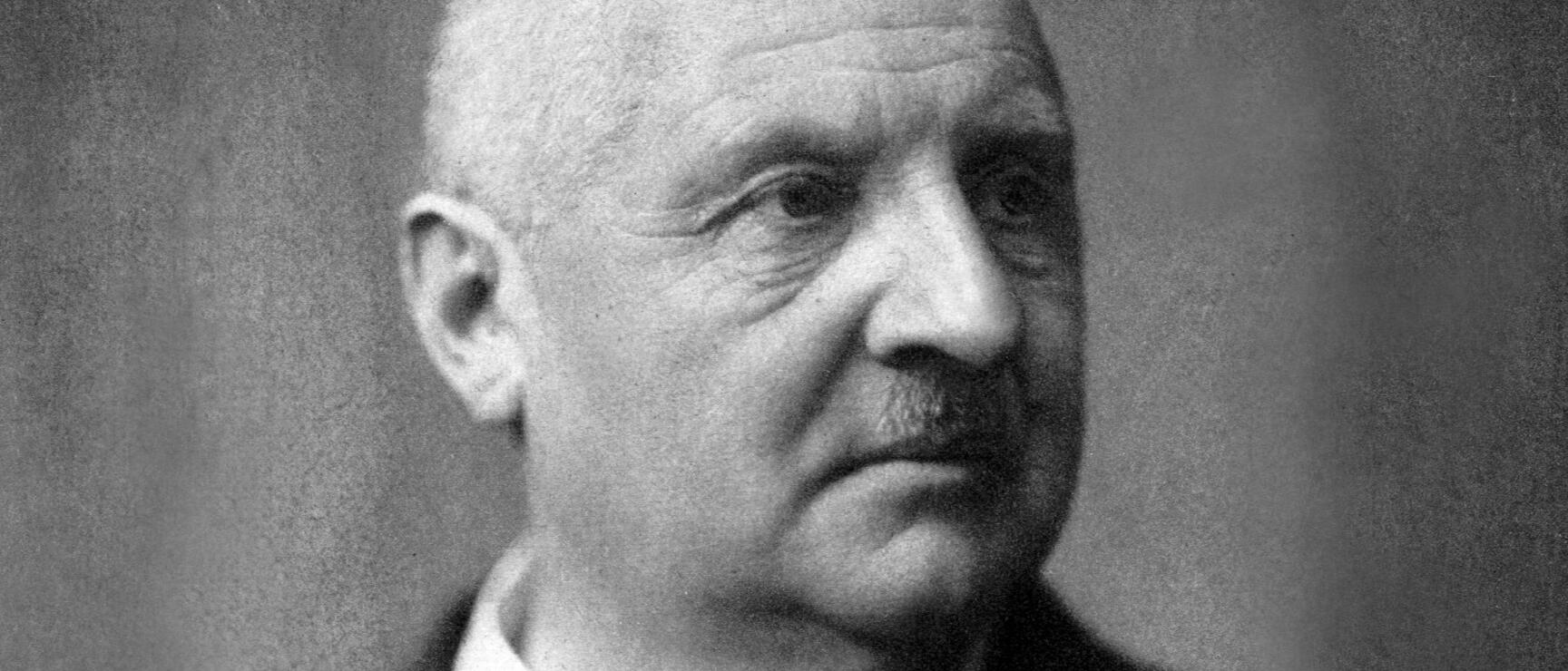 Black and white portrait of an elderly man with a bald head, small moustache and dark jacket.