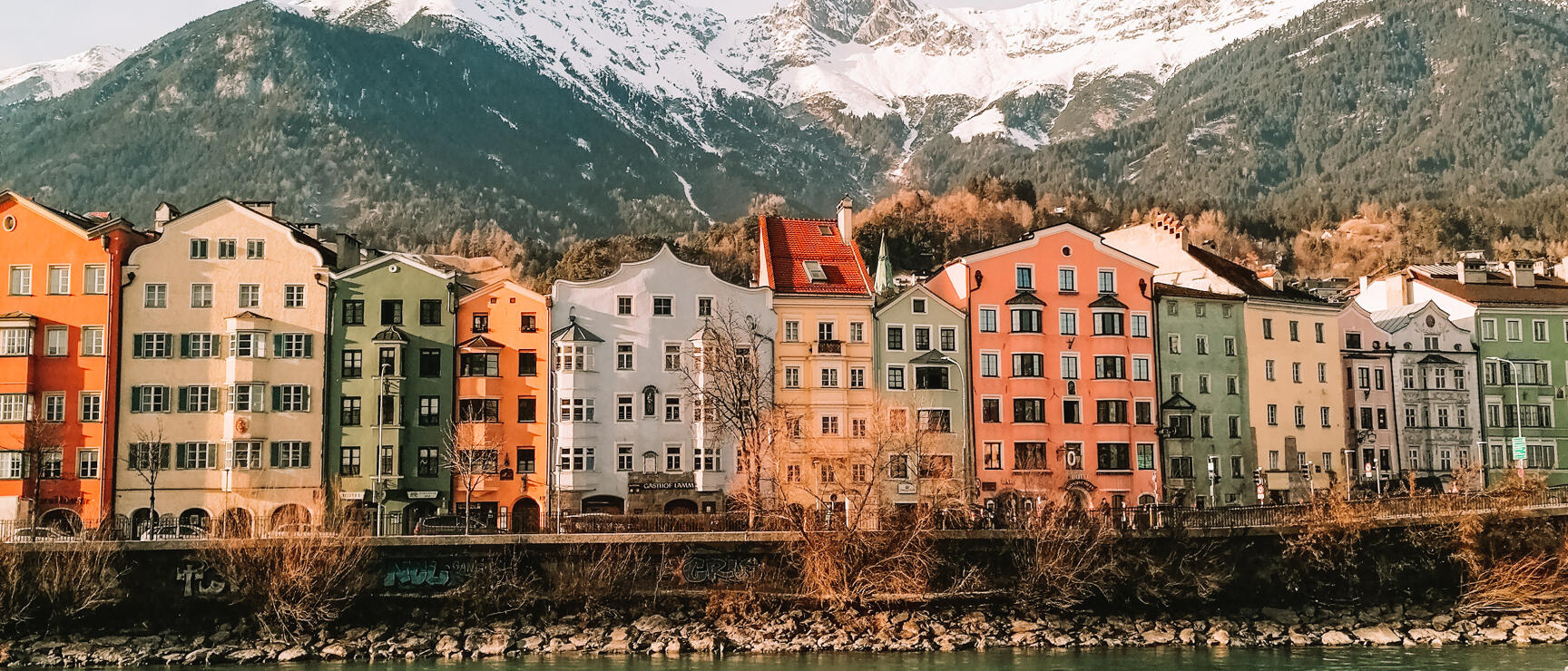 Bunte Häuserzeile am Innufer in Innsbruck, schneebedeckte Berge im Hintergrund.