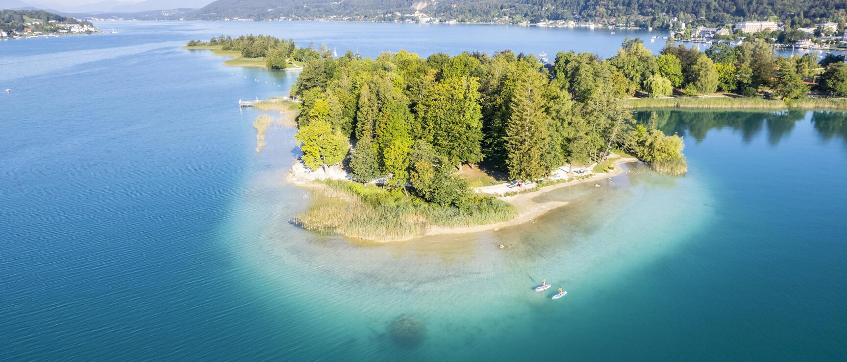 Aerial view of a forested island in Lake Wörth, two SUP paddlers in turquoise water, hills in the background.