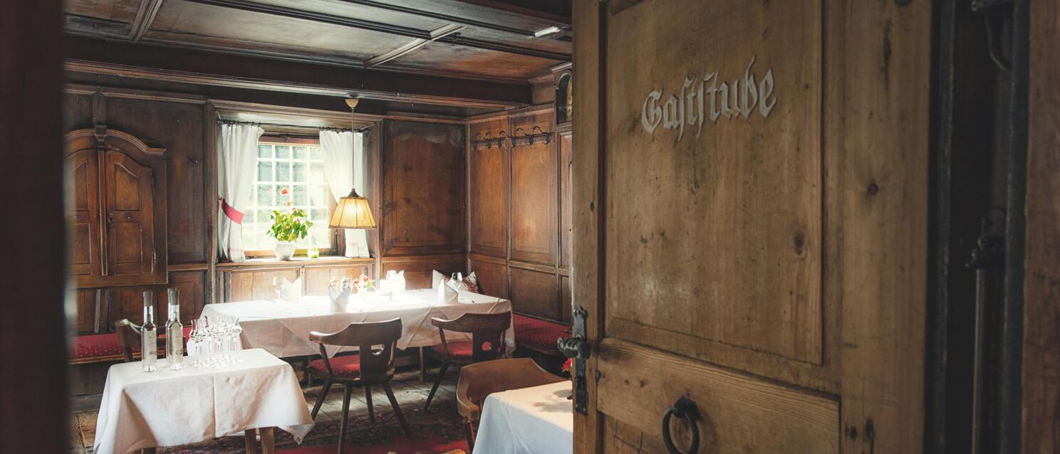 Wooden door labeled 'Gaststube', behind it a rustic pub with set tables and wooden paneling.