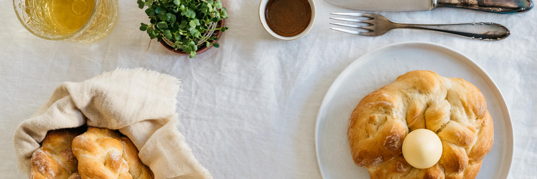 Laid table with Easter pastries, coloured egg, jam, butter, cress and cutlery on white tablecloth.