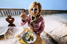Woman in sunglasses eating a salad on a snowy wooden terrace with a glass of white wine beside her.